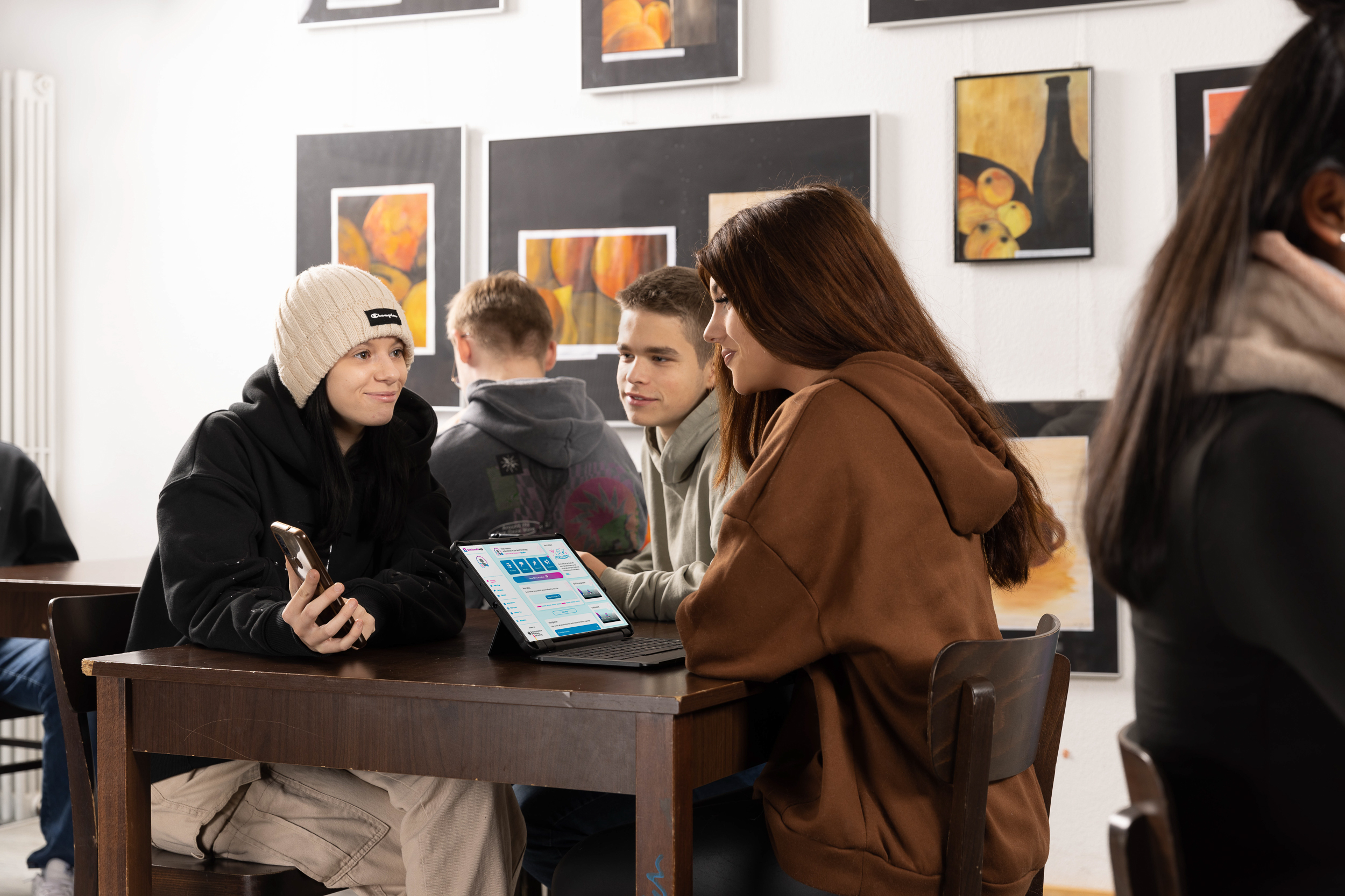 Drei Jugendliche, zwei Mädchen und ein Junge sitzen an einem Tisch im öffentlichen Raum, möglicherweise ein Café oder eine Kantine. Sie sind lässig gekleidet und unetrhalten sich. Ein Mädchen zeigt den beiden anderen Personen etwas auf ihrem Smartphone. Auf dem Tisch steht ein Laptop auf dem die berufswahlapp geöffnet zu sehen ist.