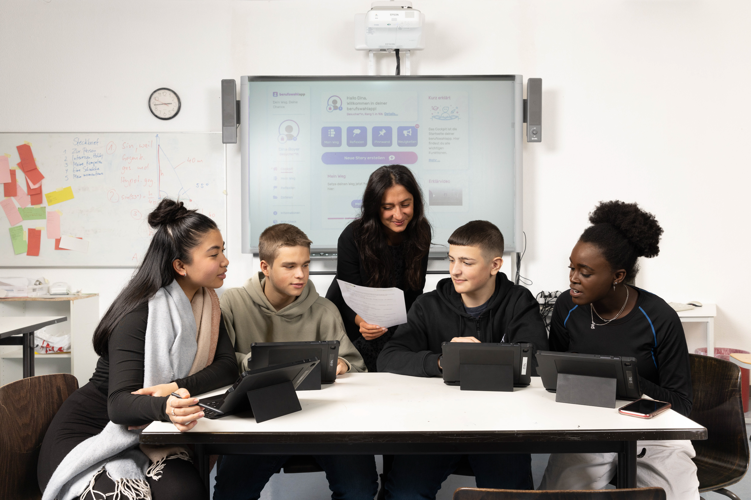 Eine Gruppe bestehend aus zwei Schülerinnen und zwei Schülern sitzt mit Tablets an einem Tisch im Klassenzimmer. Im Hintergrund wird das Backend der berufswahlapp mit einem Beamer an eine Leinwand geworfen. Die Lehrerin steht hinter den Jugendlichen und sieht ihnen bei der Arbeit zu.