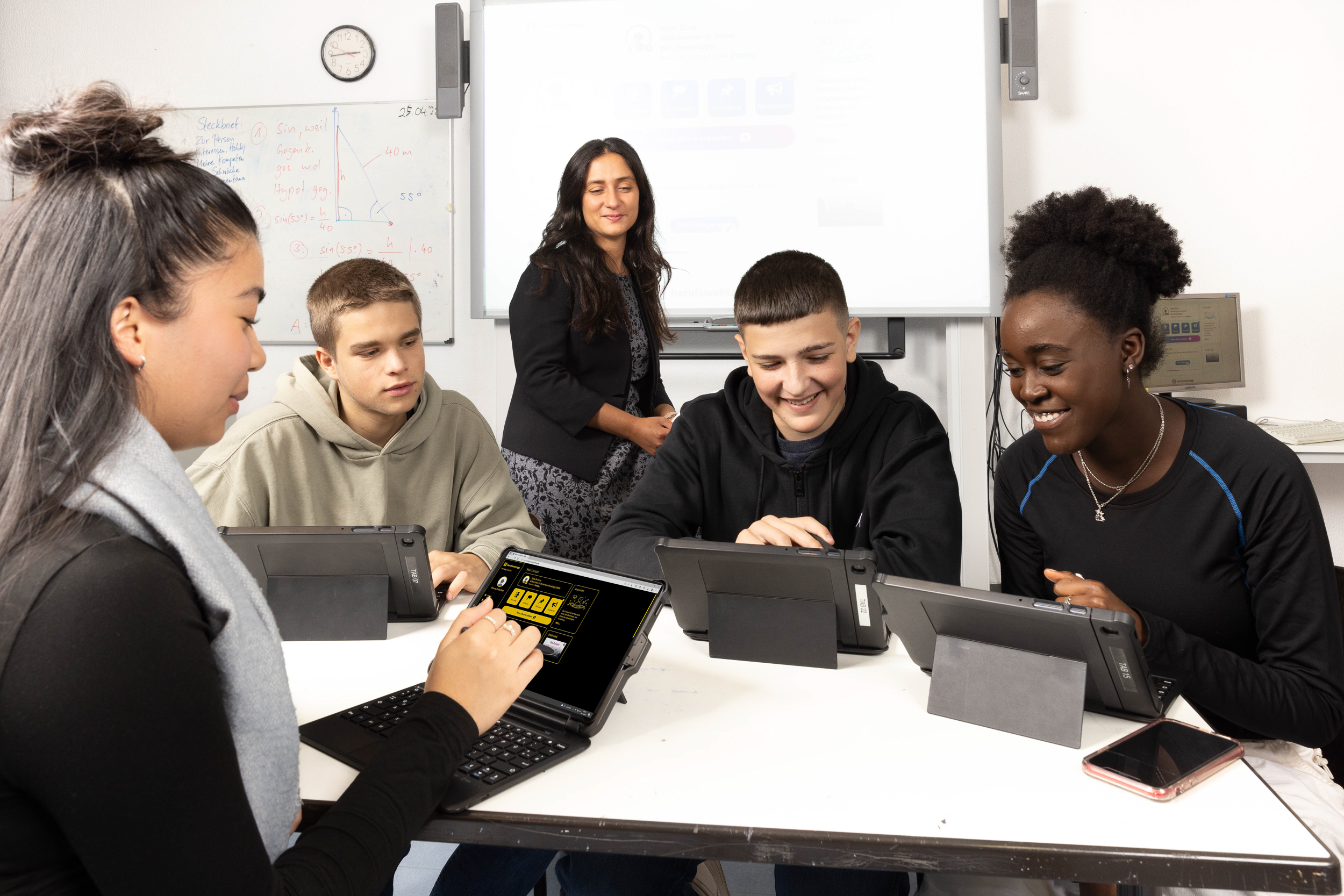 Vier jugendliche Schülerinnen und Schüler und eine weibliche Lehrkraft sitzen an einem Tisch und arbeiten mit Tablet-Computern in der berufswahlapp. Eine Schülerin im Vordergrund nutzt die berufswahlapp im Kontrastmodus