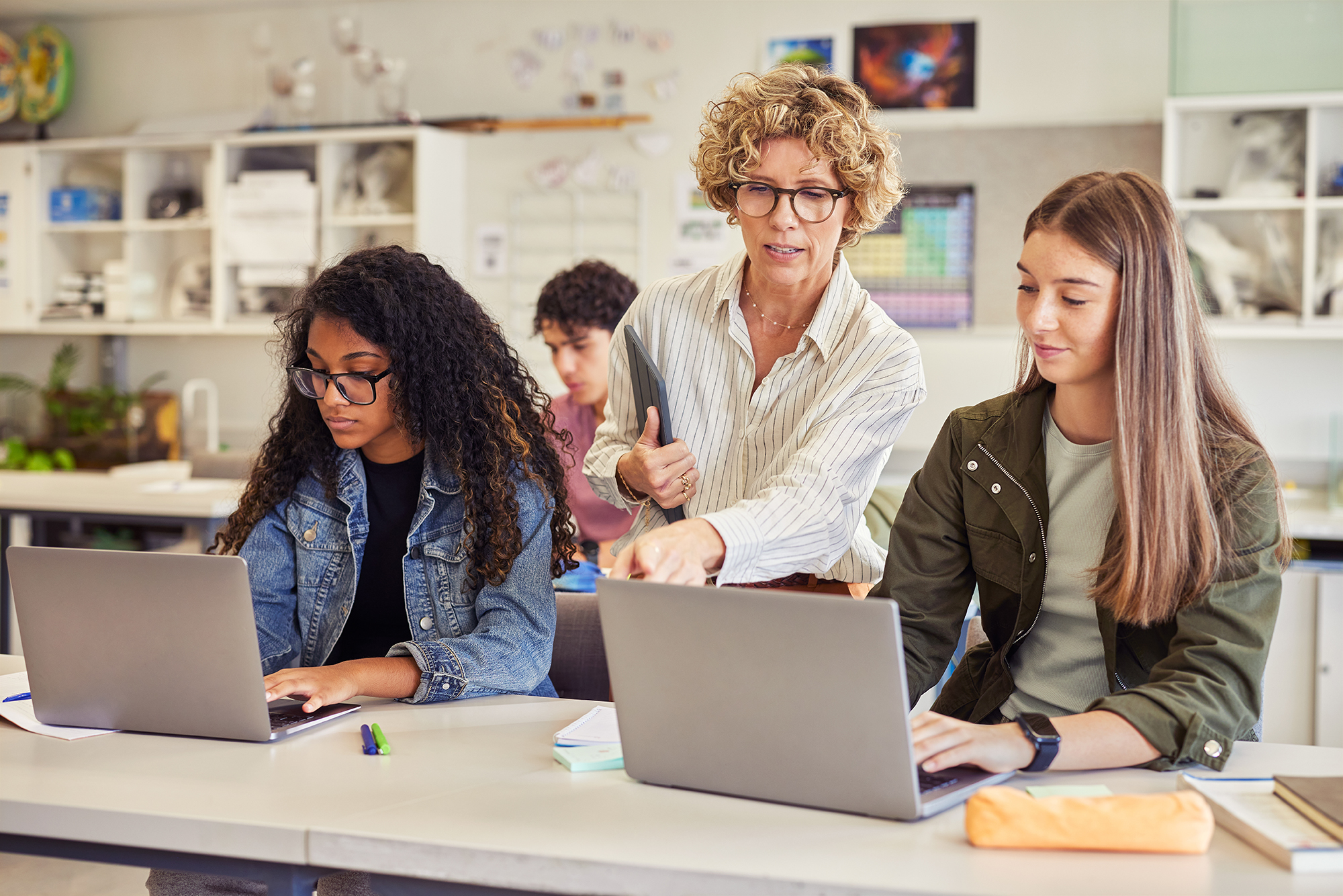 Eine Lehrkraft unterstützt zwei Jugendliche bei der Arbeit auf ihren Laptops im Klassenzimmer. Alle Personen werden weiblich gelesen.