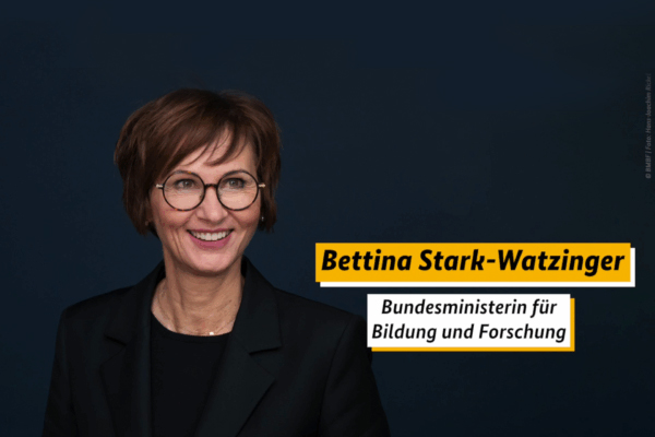 Bettina Stark-Watzinger, die zum Zeitpunkt des Artikels die Bundesministerin für Bildung und Forschung war, lächelnd vor einem dunkelblauen Hintergrund.