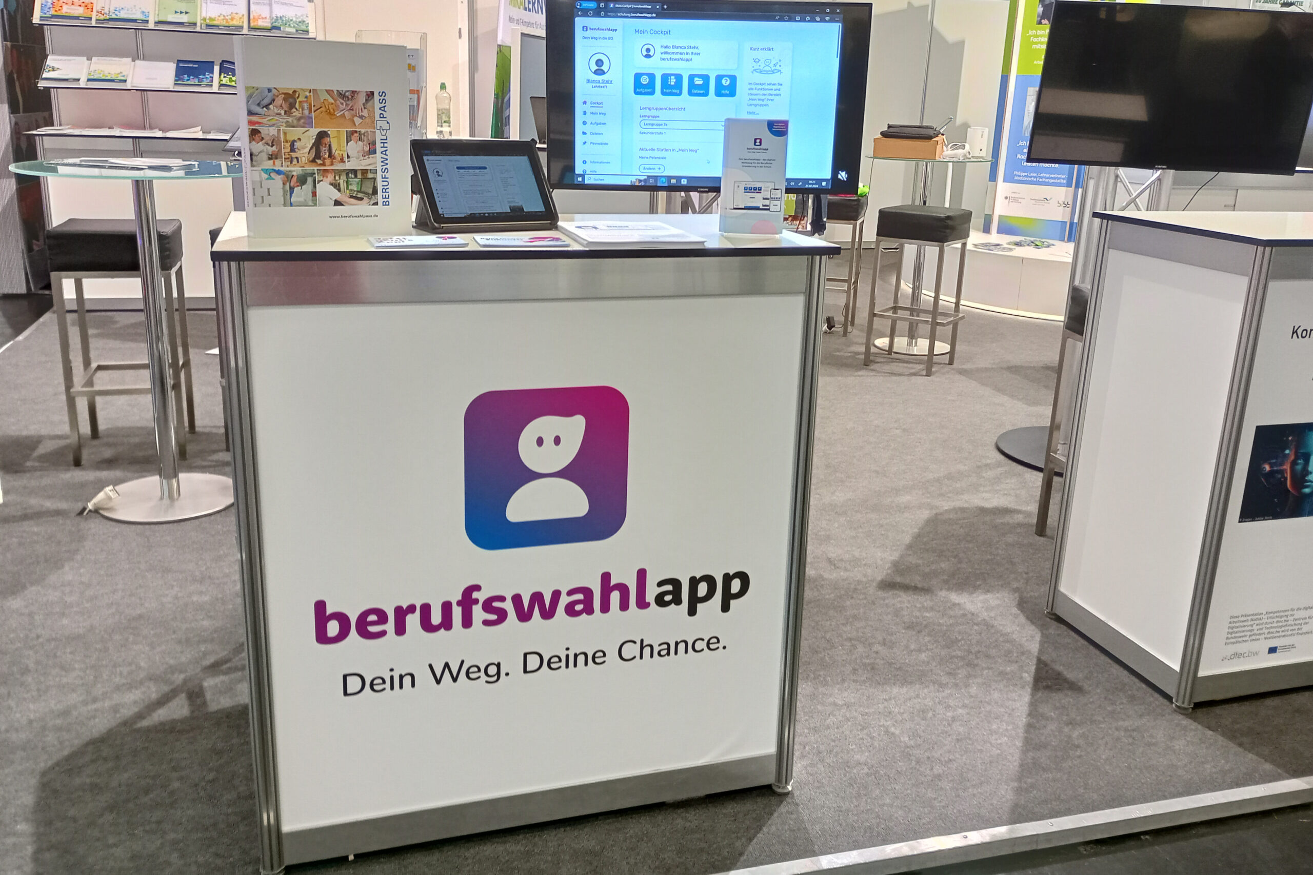 Der BIBB-Stand auf der didacta. Im Vordergrund sieht man ein großes Pult mit der Aufschrift "berufswahlapp". Darauf stehen ein Tablet, ein Ständer mit berufswahlapp-Flyern und ein Berufswahlpass. Im Hintergrund sieht man einen Monitor mit der berufswahlapp und den Rest des BIBB-Standes mit Stühlen und Tischen.